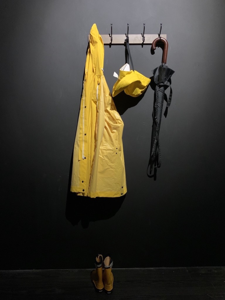 A yellow rain-mac and black umbrella with brown handle, hang from a coat hook on a black wall, both illuminated in spotlight.
