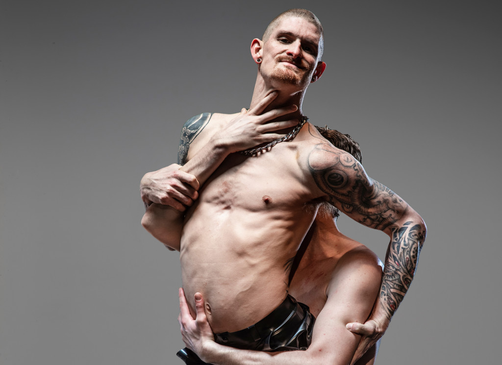 A young white man is suspended in the air, held up by another man’s arms. He is looking straight into the camera. The other man’s arms are visible around the man’s body but his face is not. The first man is topless and he has tattoos on his arms and shoulders. He is wearing black shiny trousers.