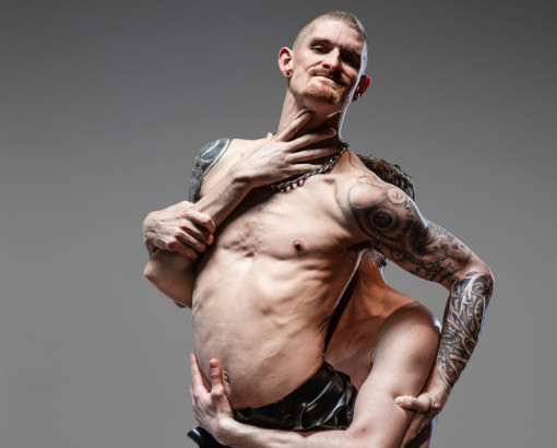 A young white man is suspended in the air, held up by another man’s arms. He is looking straight into the camera. The other man’s arms are visible around the man’s body but his face is not. The first man is topless and he has tattoos on his arms and shoulders. He is wearing black shiny trousers.