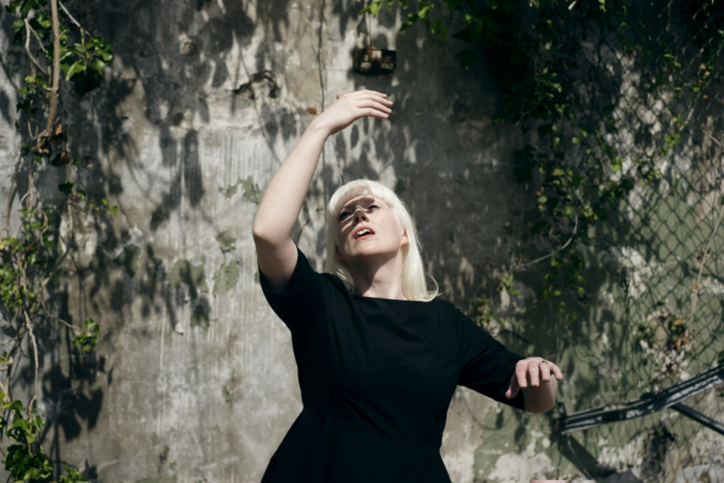 A woman with white blonde hair raises her hand above her head, casting a shadow on her face. She is wearing a black dress. She is standing outside, against a background of a grey wall with green leaves casting shadows on the wall.