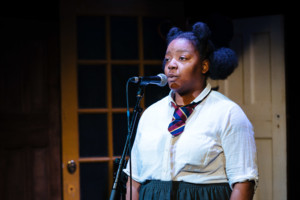 A young woman stands facing to her right. She is wearing a white shirt, grey skirt and a blue and maroon tie. Her hair is tied back and she is speaking into a microphone. In the background there are two wooden doors. 