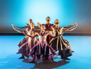 Five dancers stand on a blue stage, illuminated by light. They are squatting slightly with their arms open and poised upwards. They are wearing a mix of bright purple and grey South Indian clothing