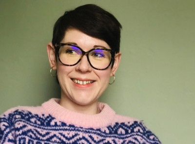 A woman with cropped dark hair and glasses looks to her right, she is wearing a pink jumper