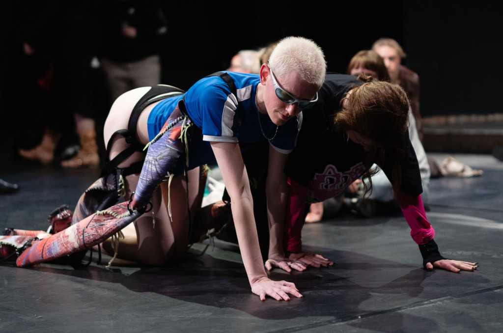 A white woman is on her hands and knees on a stage, facing towards her left. She has closely cropped bleached blonde hair, and wears sunglasses, a blue and white top, and a black thong. Another woman is beside her on her hands and knees, she has brown hair and her face is directed towards the floor.