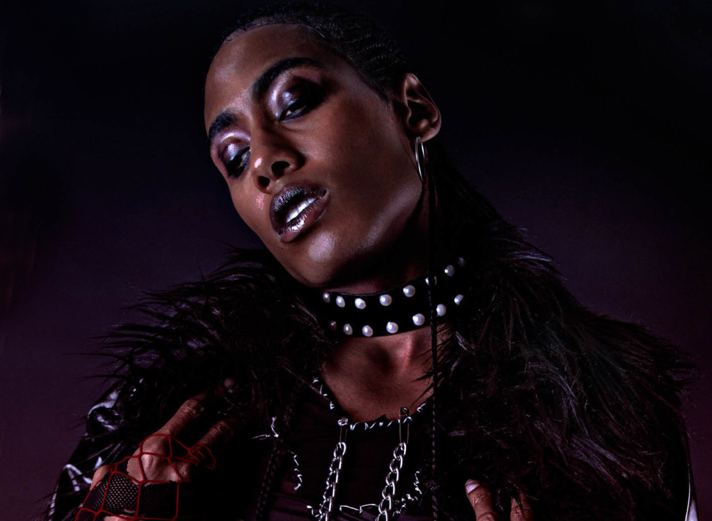 A non-binary person of the Global Majority, is standing close in frame, looking downwards to their right. They are wearing bold make-up, a black collar with white studs, silver hoop earrings and a feather trimmed coat, over a black crop top connected with safety pins and chains. They wear red and black fishnet gloves.
