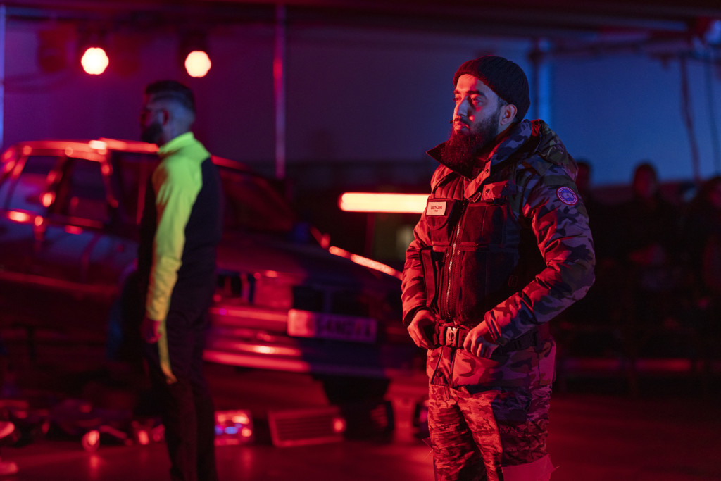 A man of the Global Majority stands facing to his right. His face is illuminated by red lighting, and he has a long beard. He is wearing a dark woollen hat and camouflage, military style clothing.