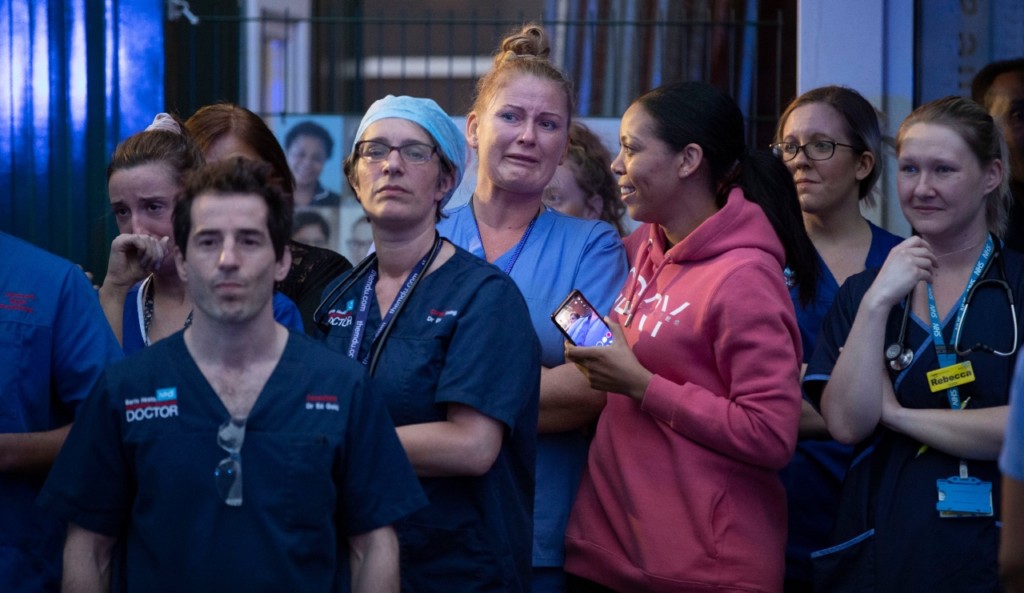 NHS workers stand facing towards the camera. They wear blue scrubs and tired expressions.