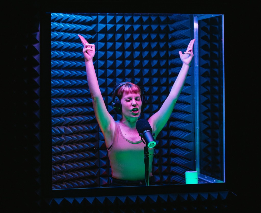 A young, white woman with short, brown hair and wearing a beige vest top stands in a recording booth. She is also wearing headphones and speaking into a large microphone, eyes closed. Her arms are thrown in the air with finger-guns pointing to the sky. She visible from the waist up and illuminated in red and green lighting. The walls of the booth are covered with spiky foam soundproofing material. Outside of the booth is completely dark.