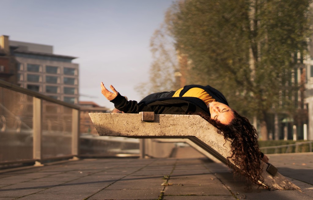 A concrete urban landscape, tower blocks, trees and metal railing set against a pale blue sky. In the foreground, a woman with long, brown, curly hair lies on her back on a concrete bench with her eyes closed. She is wearing a yellow t-shirt, a black zip-up hoodie and dark jeans. Her hair cascades over the edge of the concrete bench. Her left arm rests stretched outwards, her hand slightly lifted towards the sky.