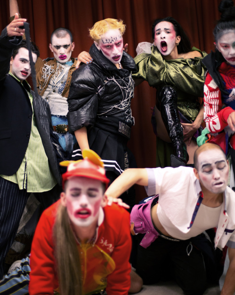 A group of seven performers wearing white facepaint with heavy blush angled upwards and bright lipstick stand in front of a red curtain. Their costumes are an assortment of colourful, thrifted, cropped, stitched together, with shiny and crinkly textures. Two performers are on all fours in the foreground, slightly out of focus, eyes closed, mouths open. The remaining five performers stand in the background, looking directly at the audience, some scowling, one with her mouth wide open, one smiling slightly.