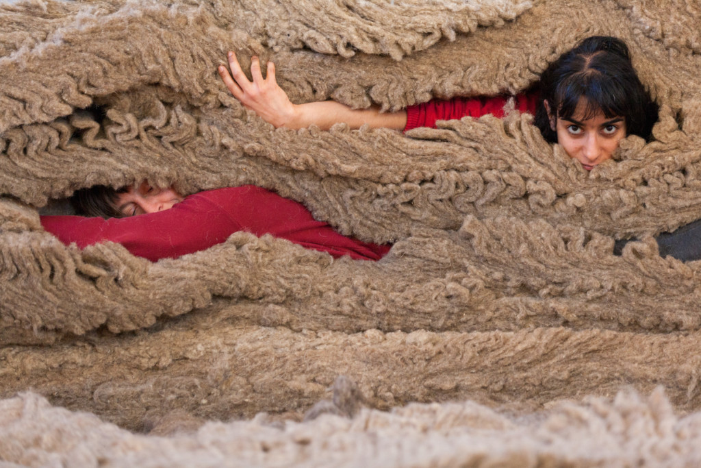 Out from ten layers of dusty beige sheeps wool peek two women in red tops, only their faces and one arm visible. On the right, a Middle Eastern woman with black hair and a fringe looks directly at us with one arm reaching out to the left and her hand loosely grips the wool. Underneath her and to the left is an older woman with grey hair lying on her side, head resting on her arm, eyes closed as if asleep.