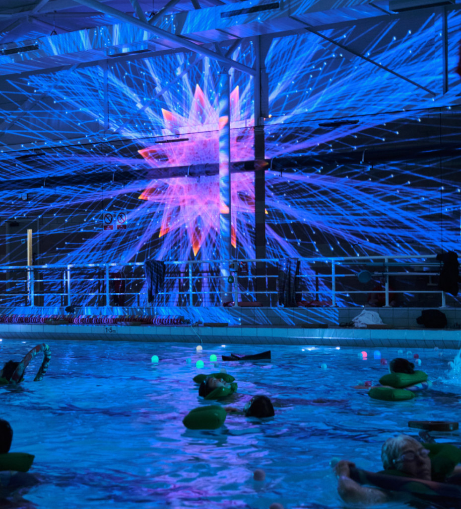 A dark swimming pool is illuminated by a kaleidoscopic blue and pink wall projection, which makes the space glow blue. A few swimmers recline in the water, using pool noodles to keep them afloat. Small orbs glowing green, pink, and blue float in the water with them.