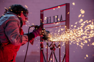 A Jewish woman with dark brown curly hair is welding an iron gate. She is wearing a distressed denim shirt and a brown leather apron as well as protective goggles and gloves. Sparks are flying from her welding tool. On the iron gates stands the word "AFTER" as seen from the back in reverse on the top section. The scene is washed in a traquil pale purple shade juxtaposed against the red hot glow of the welding, which illuminates the woman and the gate.