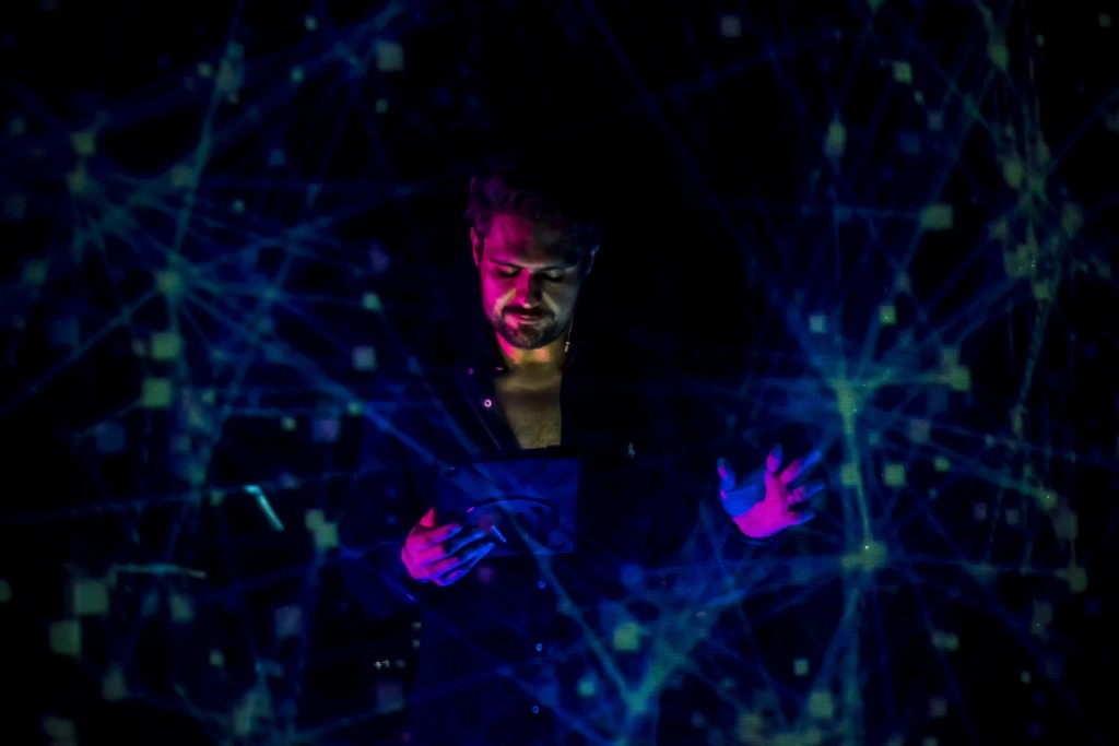 A Middle Eastern man with short, dark curly hair and a dark beard, wearing a dark shirt unbuttoned to reveal the top part of his chest stands in a dark space. The darkness is illuminated by neon green dots and intersecting connecting lines in a busy network structure surrounding him. He is looking down at an iPad, his hands and face illuminated with neon pink light. His left hand is outstreched, reaching towards the green network.