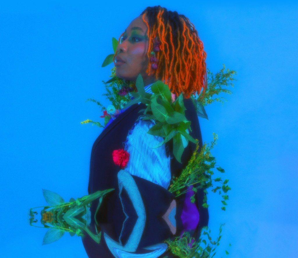 Against a vibrant blue background a Black performer stands side-on facing the left. They wear their hair in vibrant orange twists. They are wearing a loose fitting striped pale blue long sleeved shirt and a navy waistcoat with a red flower in the breast pocket. Flowers and leaves appear to grow out of them and their clothing. Their head is tilted slightly upwards, looking on with a neutral expression. In the bottom half of the image we see an upside down rippled reflection, as if it's a reflection on the water's surface.