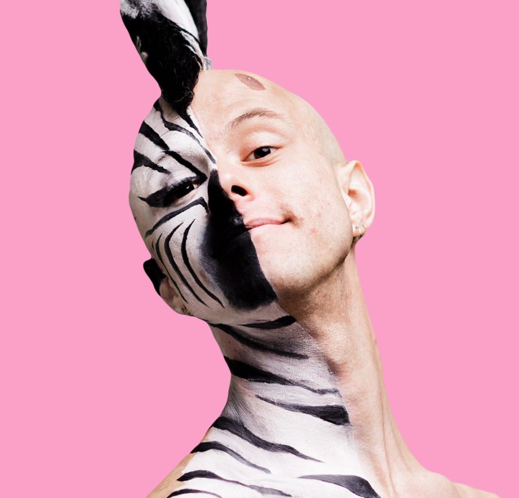 Against a bubblegum pink background stands a young, white, bald man, visible from the chest upwards. He is wearing nothing on his top half. His head is held high and tilted to the left and he is smiling. He is half painted down to his collar bone with black and white facepaint as a fabulous zebra with a black and white mohawk and false lashes. The other half is bare apart from a brown birthmark above his eyebrow.