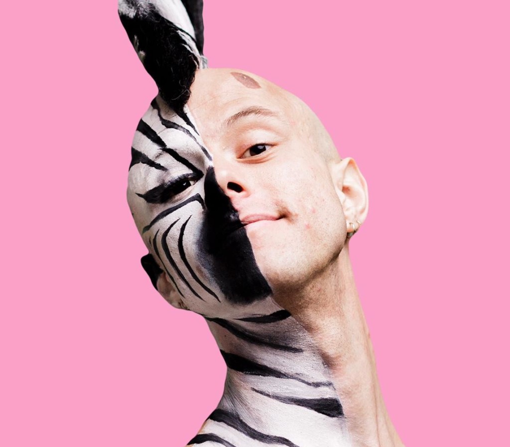 Against a bubblegum pink background stands a young, white, bald man, visible from the chest upwards. He is wearing nothing on his top half. His head is held high and tilted to the left and he is smiling. He is half painted down to his collar bone with black and white facepaint as a fabulous zebra with a black and white mohawk and false lashes. The other half is bare apart from a brown birthmark above his eyebrow.