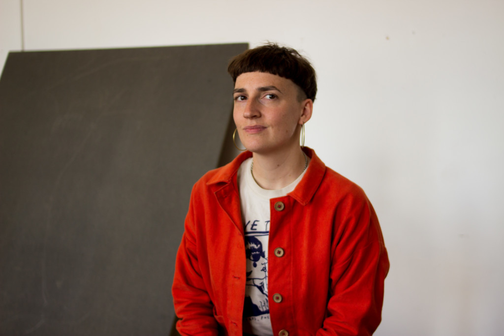 A white, non-binary individual sits visible from the waist up. They have cropped brown hair and are wearing large gold hoop earrings and a red shirt with wooden buttons over a white t-shirt. Behind them is an out of focus dark grey slab, leaning against a white wall.