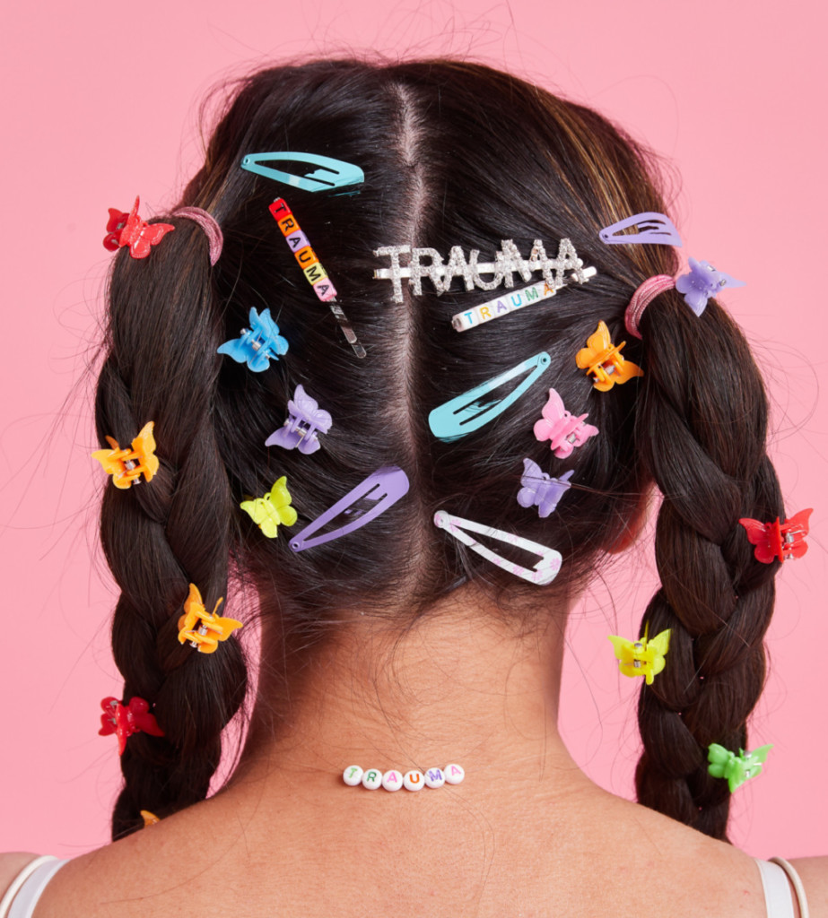 Back of a South Asian woman’s head against a candyfloss pink background. Black hair is covered in early 2000s inspired hair slides, butterfly clips. One silver hair clip reads the word ‘TRAUMA’. White plastic beads stuck to the back of the woman's neck also spell out the word 'TRAUMA'.