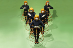 The camera looks down on six white women standing against a bright green background in a V formation, looking up. They are wearing, bright orange wigs in a bob hairstyle, striking orange toned stage makeup, full body black leotards, bright orange hoop cage skirts, and bright orange and black trainers.