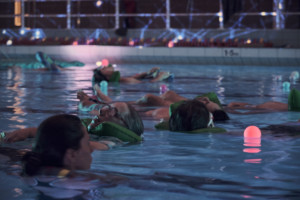 In a swimming pool, participants float with green headrests. The lighting is dim and atmospheric, with pink orbs floating in the water and a kaleidoscopic image projected on the wall behind the pool.