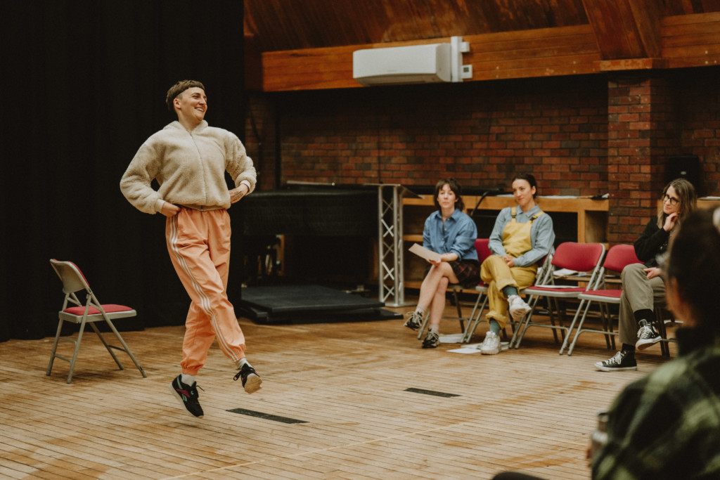 In a rehearsal room with wooden floors and bare brick walls, a white performer wearing a beige fleece, orange tracksuit bottoms and black trainers jumps straight up in the air with their hands on their hips and one leg raised slightly in front of the other. They have cropped brown hair. Sitting around them and observing in red chairs to the right are four white women.