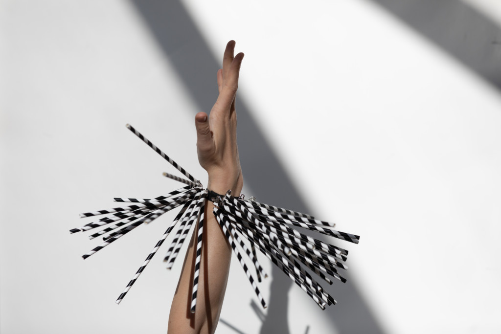 Against a white background with two shadow lines, a hand with white skin reaches up, wearing a bracelet with long black and white stripy tassles hanging off it and swinging in motion.